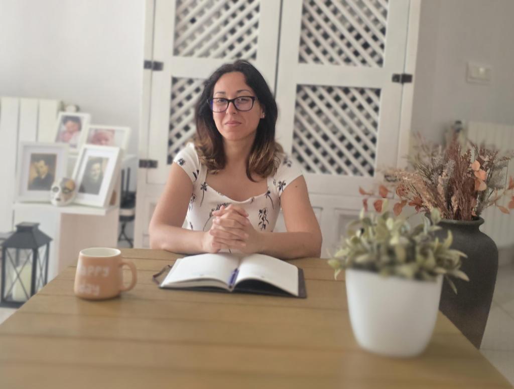 Founder Sonia Perez with glasses sitting at a wooden table, hands clasped, looking at the camera. An open notebook and a coffee mug are in front of her. The background features decorative items and plants.