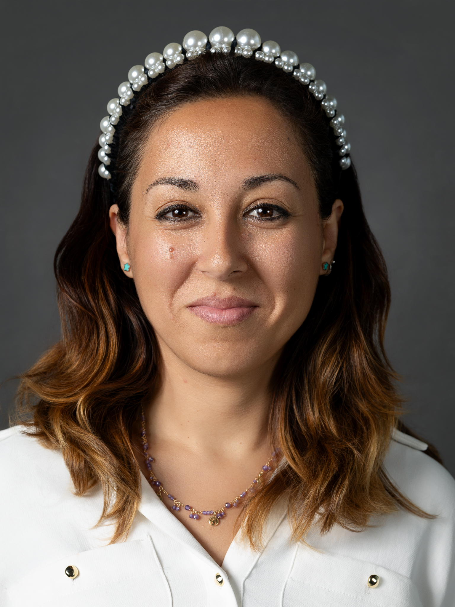 Portrait of founder Sonia Perez with long brown hair wearing a pearl headband and a white blouse, smiling softly at the camera against a dark background.