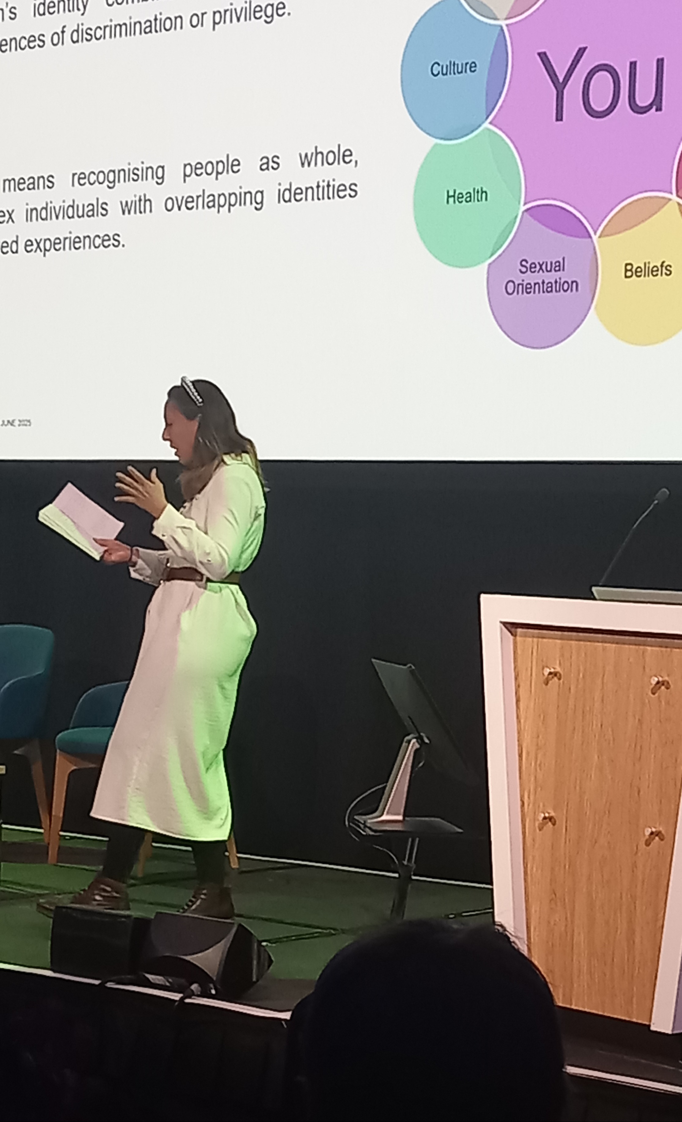 Founder Sonia Perez in a white dress presents on stage, holding notes, with a diagram on a screen behind her summarizing themes of identity, culture, health, and beliefs.